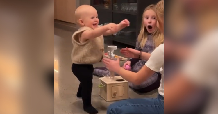 Baby boy smiles wide as he takes his first steps. He has his arms out reaching for one of his sisters who is reaching out for him, too. His second sister sits nearby, mouth open from shock as she looks over at the person recording this moment.