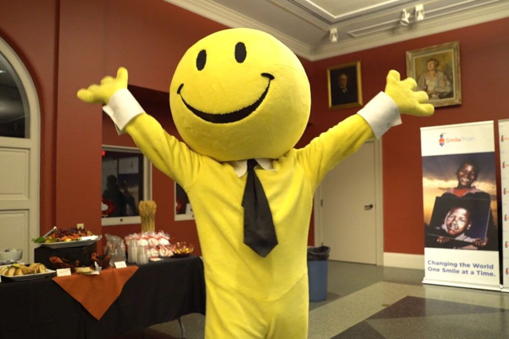 Someone dressed in a yellow onesie with a tie and a full-face mask of the iconic yellow smiley face raises their hands in celebration.