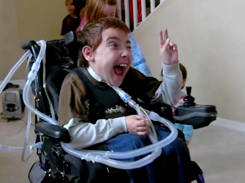Jacob smiles wide as he gives a peace sign. He's in a wheelchair with several tubes connected to him.
