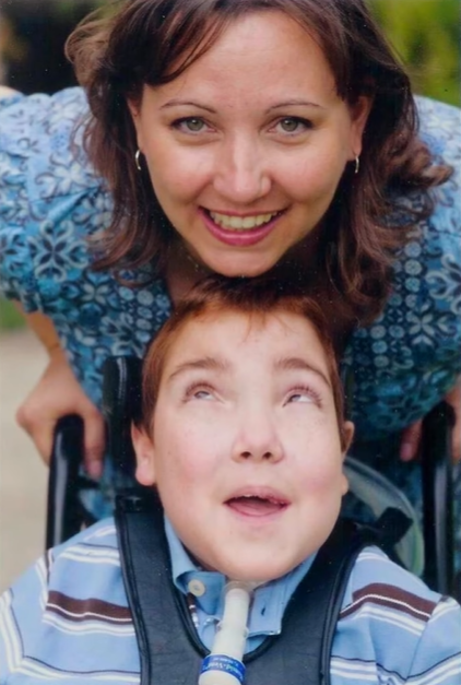 Jacob, who is sitting in a wheel chair, smiles as he looks up at Stacy who is smiling at the camera.