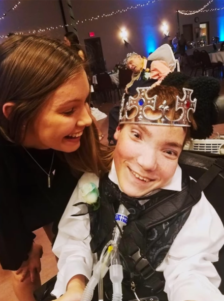 Jacob smiles from his wheelchair as he wears a crown at Night to Shine. A woman smiles as she looks at him and leans in to be closer to him.