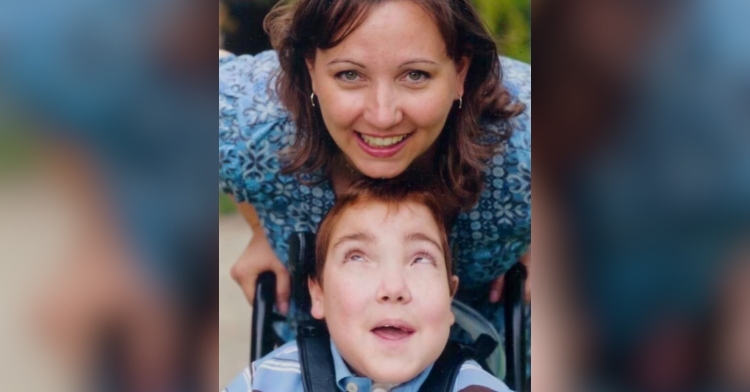 Jacob, who is sitting in a wheel chair, smiles as he looks up at Stacy who is smiling at the camera.