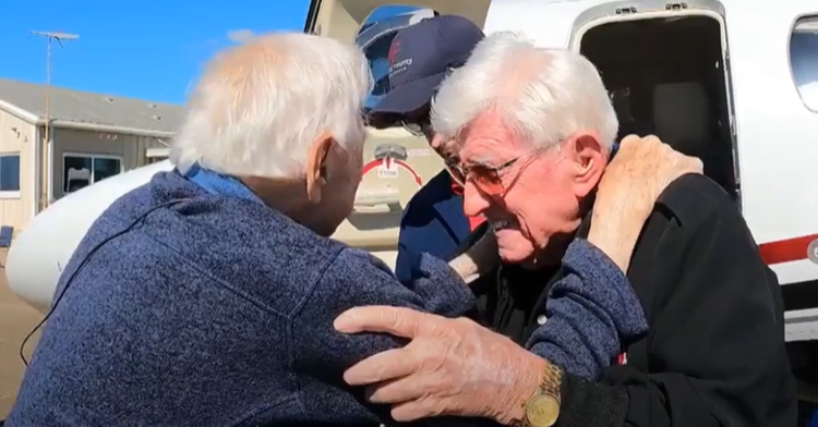 Two elderly men who are both veterans have their hands on each others shoulders as they start to hug each other. We can see one of their faces and he looks emotional.
