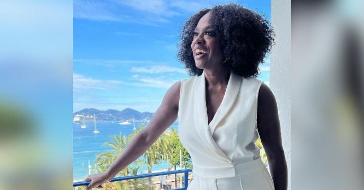 Viola Davis smiles as she looks in the distance and stands on a balcony, a beautiful ocean in the distance behind her.