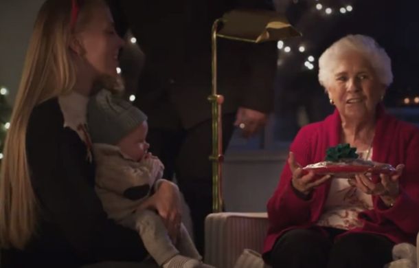 Image shows a young woman with a baby visiting an elderly person, who is holding a wrapped tray containing Christmas goodies.