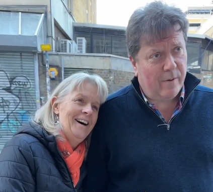 A woman named Sue smiles, mouth open as she rests her head on her husband's shoulder. Her husband, Alex, is talking to someone we can't see in the image.