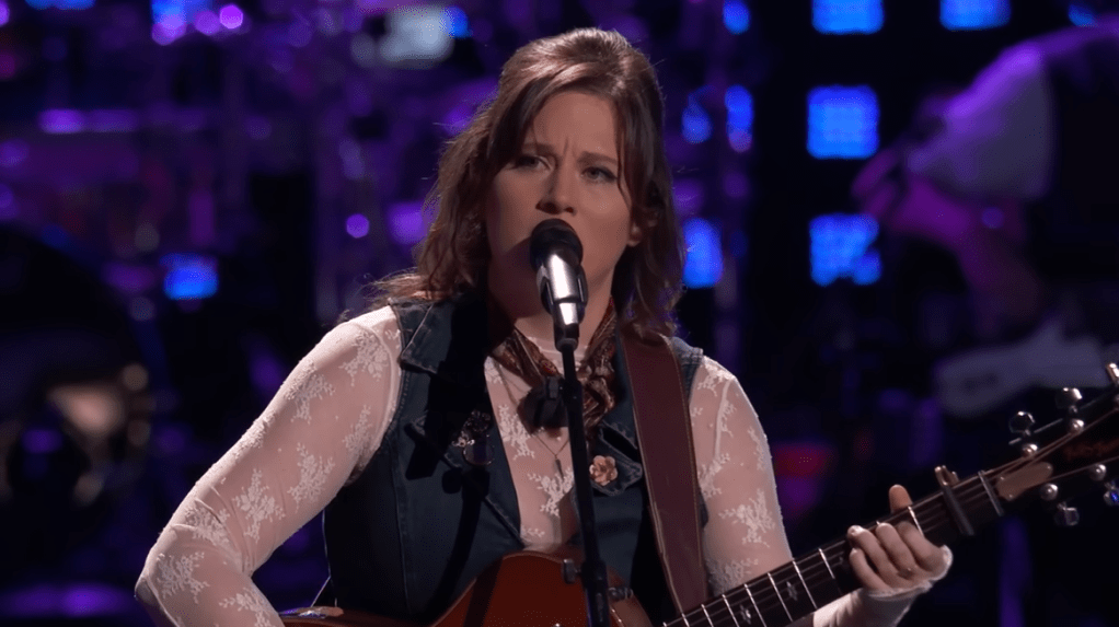 Alexa Wildish looks into the distance as she sings into a mic and plays the guitar on "The Voice" stage.