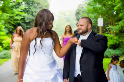 A black woman in a wedding dress smiles as she looks over at her white groom. He is also smiling as they fist bump.