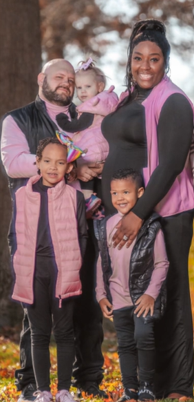 A black woman and her white husband smile as they pose with their three kids in a wooded area. The two older kids, who are standing, have darker complexions, more similar to Mom. The baby in Dad's arms has white skin like him.