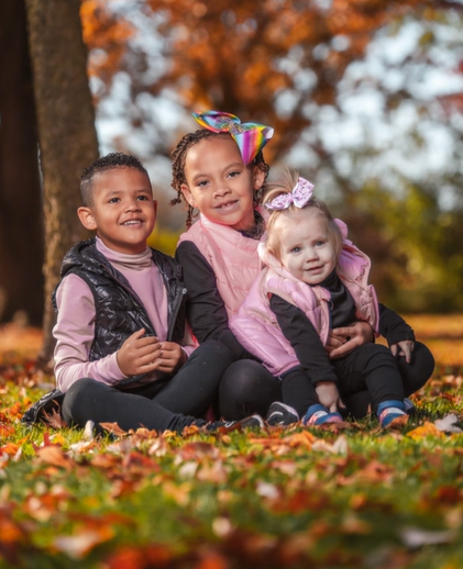 Three little kids sit on the ground in a forest area, smiling. One is a baby, and she's sitting on the eldest's lap.