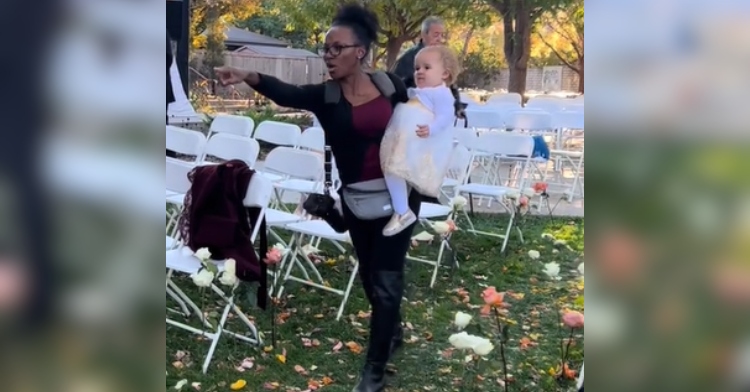 A woman who is a wedding photographer points as she holds a little girl (the flower girl) in her arms. They're standing outside. White chairs are lined up behind them and there are flower petals on the ground.