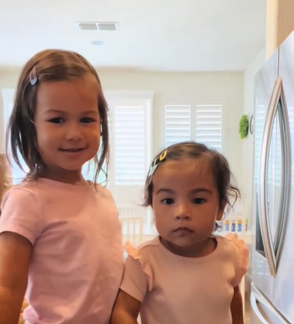 A 3-year-old and a two-year old standing next to each other, wearing the same peach-colored shirt. The older one has a smile on her face while the younger one stares blankly.