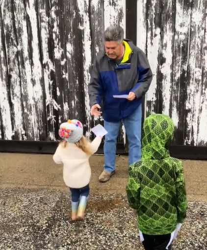 A man leans down as a little girl hands him a piece of paper. Another child stands nearby. They're outside.