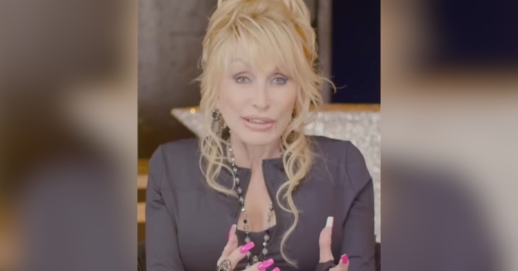 Dolly Parton gives a small smile as she talks. She uses her hands some as she talks, showing off her long, bright pink nails.