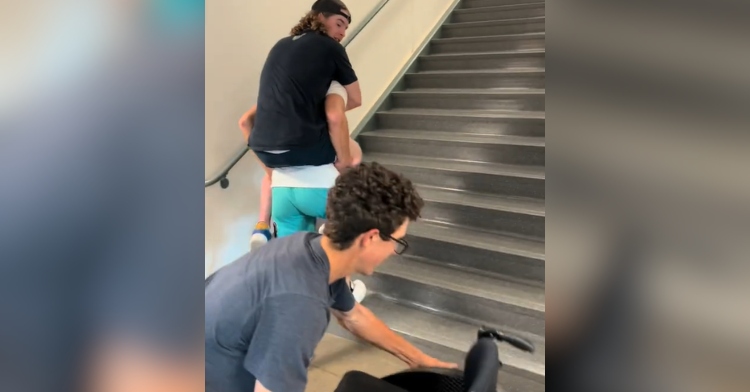 A man carrying a paralyzed man starts to walk up the stairs. The paralyzed man is looking back at a man who is picking up his wheelchair.