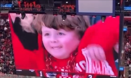 A 4-year-old named George grins as his mom lifts up one of his arms to cheer while he's being shown on the jumbotron at a Detroit Red Wings hockey game.