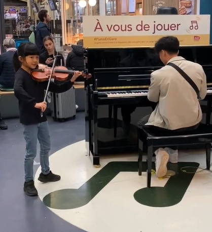 Inside a train station, an 8-year-old girl shyly plays the violin next to a man playing a piano.
