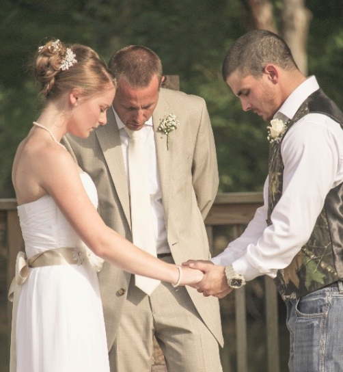 A bride and groom hold hands at the alter as they close their eyes, praying. The officiant also has his eyes closed, praying. The ceremony is taking place outdoors, and the groom's vest is camo.