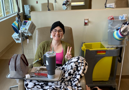 Chelsea smiles and gives a peace sign as she sits in a medical facility getting chemotherapy.