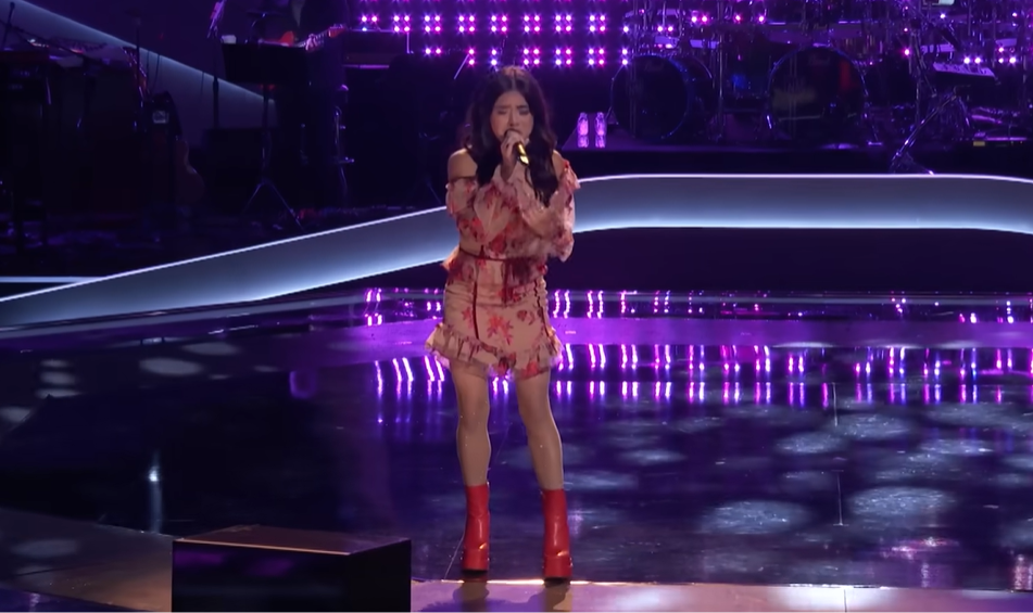 Kaylee Shimizu stands on "The Voice" stage in a red and pink dress and red boots, singing into the mic with her eyes closed.