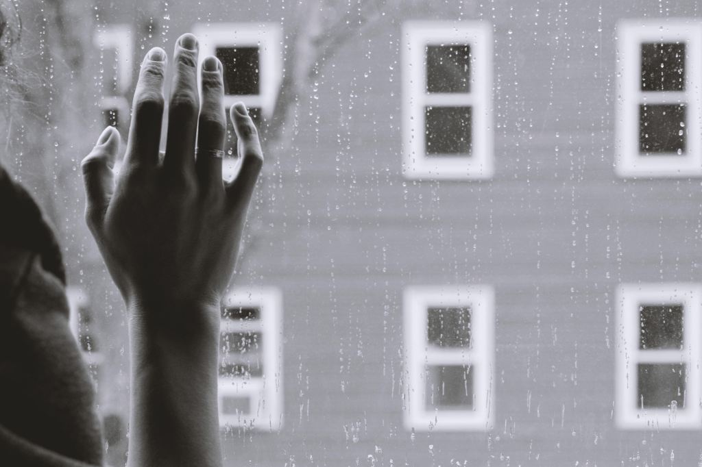 Black and white photo of a woman's hand lightly pressed against a window with rain falling down it. The view is of a building with lots of small windows.