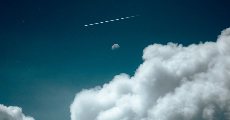 View of large, fluffy clouds just below the Moon, which is at a distance. Above the moon is a "shooting star," or rather, a meteor.