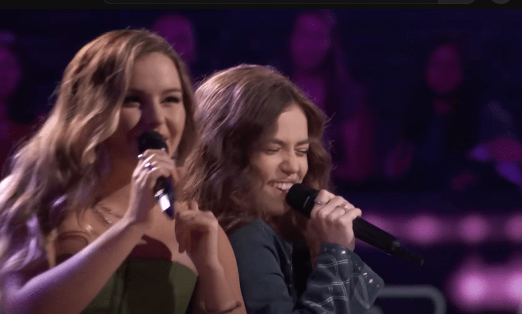 Claudia and Mara stand back to back as they sing on "The Voice." Both of them smile as they sing and Mara has her eyes closed.