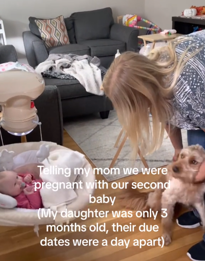 A woman leans over to pet a dog. In front of her is a baby in a bouncer, smiling. Text on the image reads: Telling my mom we were pregnant with our second baby (My daughter was only 3 months old, their due dates were a day apart)
