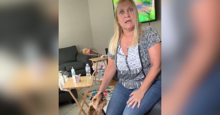 Woman sitting on the edge of a couch looks at the person taking this image, eyes wide with shock. With one hand she's petting a dog that's sitting on the floor.