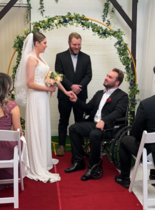 View of a wedding altar. The groom, who is in a wheelchair, smiles up at his bride as they hold hands. The officiant also smiles.