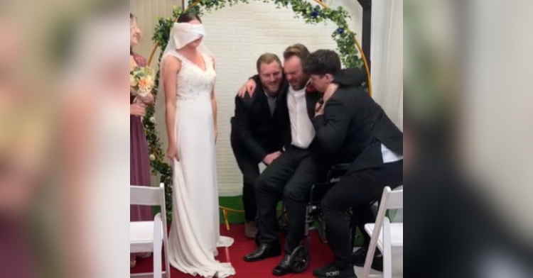 View of a wedding altar. Two men assist a man in a wheelchair onto his feet. The bride is standing next to them, a blindfold covering her eyes.