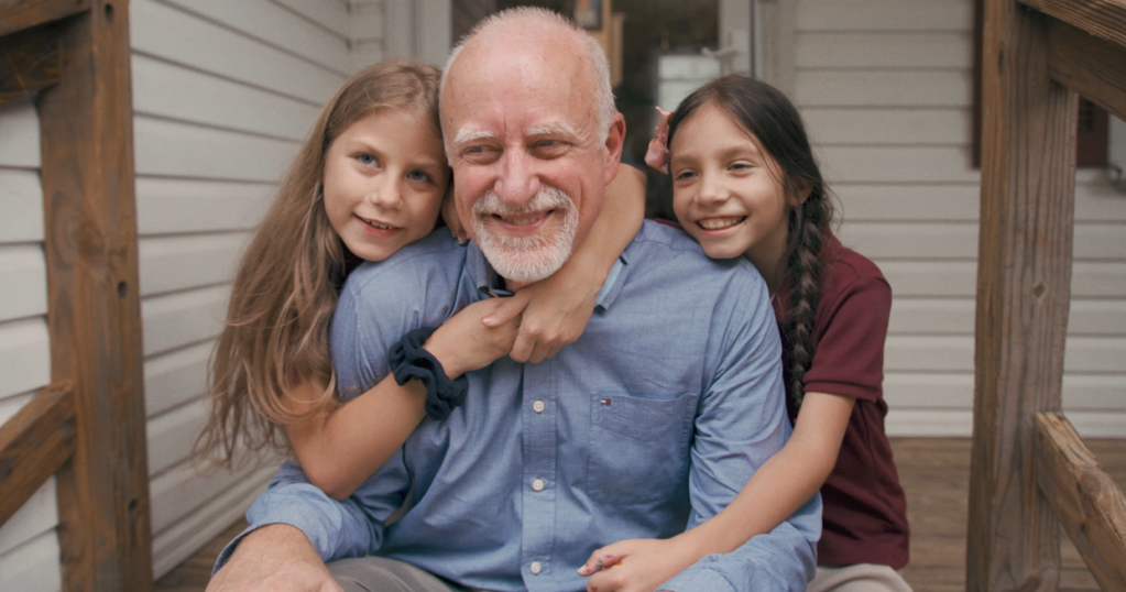 A man smiles as he sits on a front porch step with two little girls. Both are hugging him and smiling.