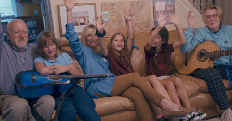 All six members of the Petrich family sit on a couch. They have their hands in the air with excitement and are smiling.
