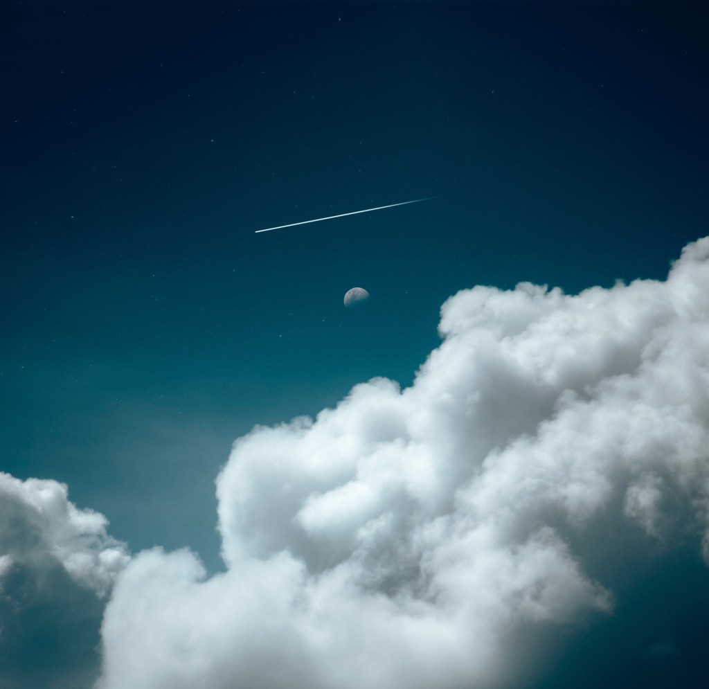 View of large, fluffy clouds just below the Moon, which is at a distance. Above the moon is a "shooting star," or rather, a meteor.