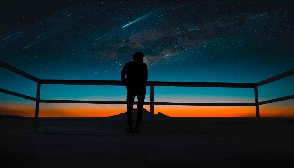 View from behind of someone leaning on railing outside as they look at the sky that is lit up by a meteor shower. Because it's dark, we only see a silhouette of the person.