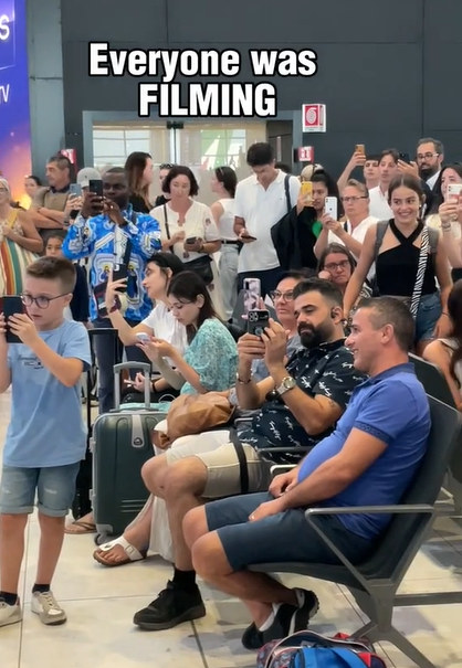 A large group of people in an airport; some are sitting and some are standing. Many of them have their phones out and are recording something taking place outside of the image. Text on the image reads: Everyone was FILMING 