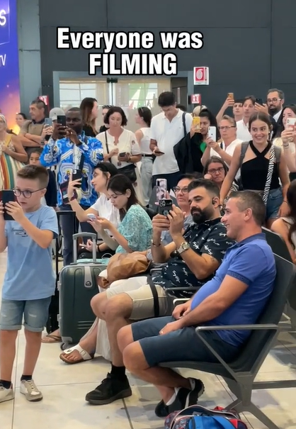 A large group of people in an airport; some are sitting and some are standing. Many of them have their phones out and are recording something taking place outside of the image. Text on the image reads: Everyone was FILMING