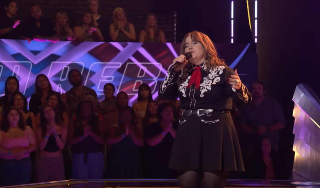 Ruby Leigh sings with her eyes closed as she performs on "The Voice" stage. The audience can be seen behind her, happily watching as they clap for her.