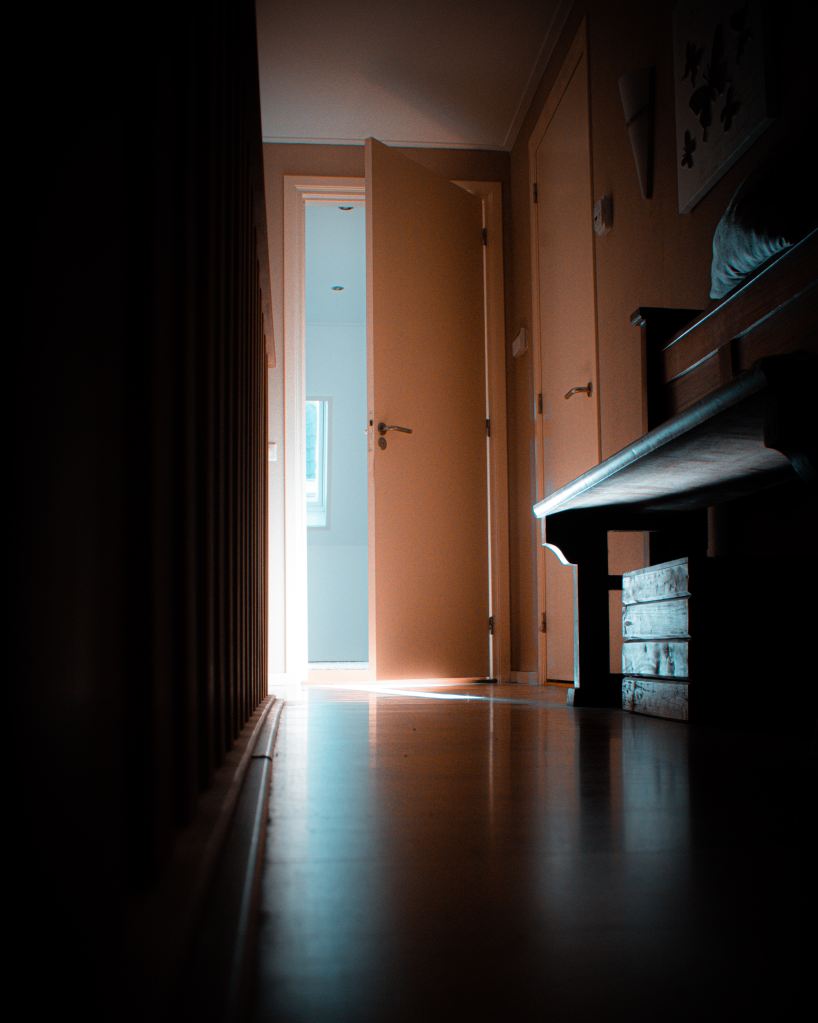 View in a dark hallway. A single door is open and is letting light inside.