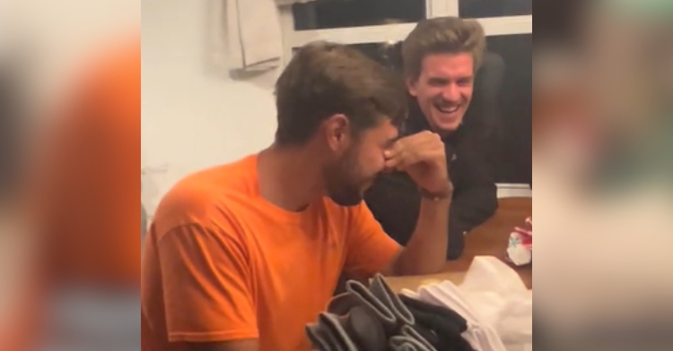 A young man sits at a table, rubbing his eyes with his hands. Another guy sits next to him, laughing.