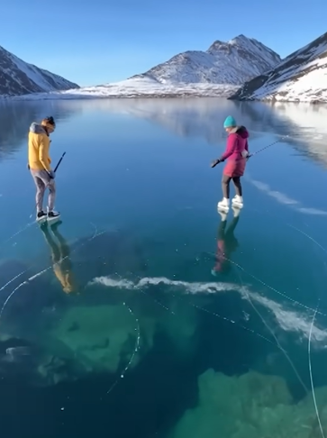 Two ice skaters stand on a frozen lake in Alaska that is super clear. Snowy mountains can be seen in the distance.