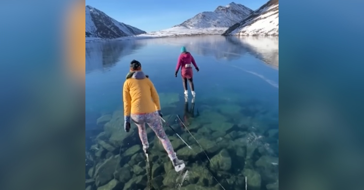 View from behind of two people ice skating on a super clear frozen lake. Snowy mountains can be seen in the distance, and the sky is clear and super blue.