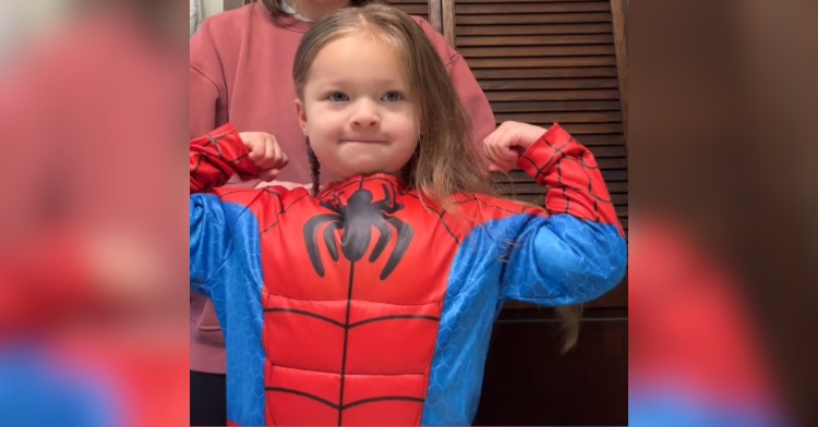 Close up of a little girl flexing her "muscles" while wearing a Spider-Man costume. She's looking at herself in a mirror.