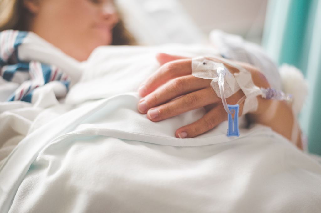 View of a woman laying in a hospital bed with a focus on her hand which is hooked up to an IV.