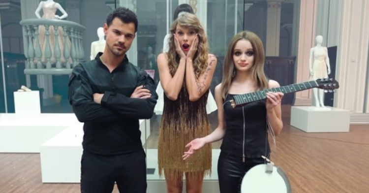 Taylor Swift places her hands on her face, pretending to look surprised. On one side is Taylor Lautner who is posing seriously with his arms crossed. On the other side of Swift is actor Joey King who shrugs as she holds a broken banjo.