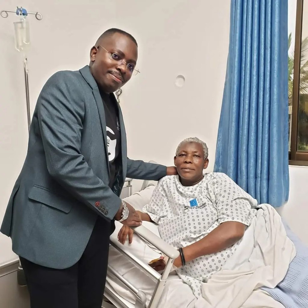 Safina, the 70-year-old who gave birth to twins, smiles from a hospital bed as she poses with a young man who stands next to her, smiling as he rests a hand on her shoulder and the other on her wrist. 