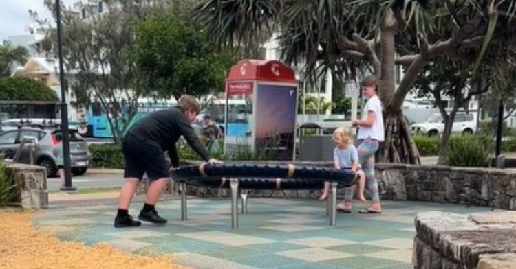 A teen boy at a park slowly spins a young child on playground equipment.