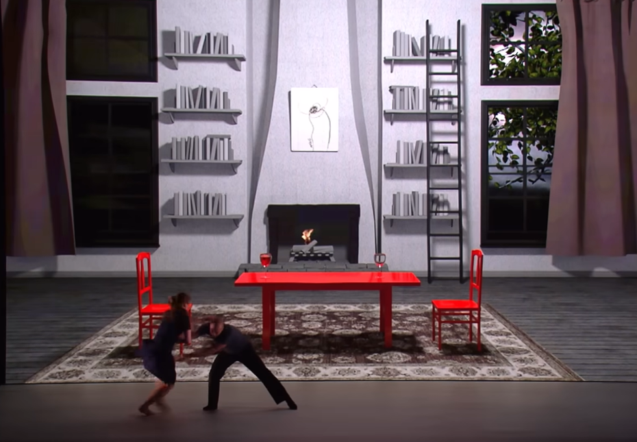 A woman and man dance in front of a screen that displays a dining room space with a fireplace and ladder in the distance.