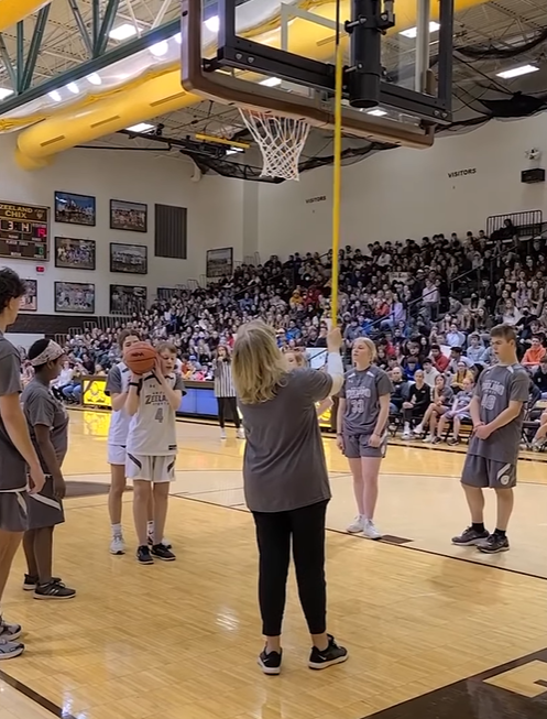 Someone assists a blind teen as she prepares for her free throw. A woman stands behind the hoops, using a stick to make sound. 
