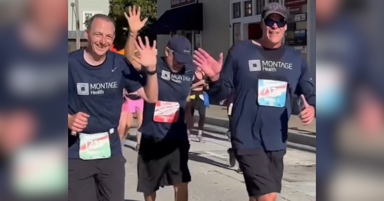 Three men running in a marathon together wave at the camera. One of them is looking down while the other two look at the camera and smile.